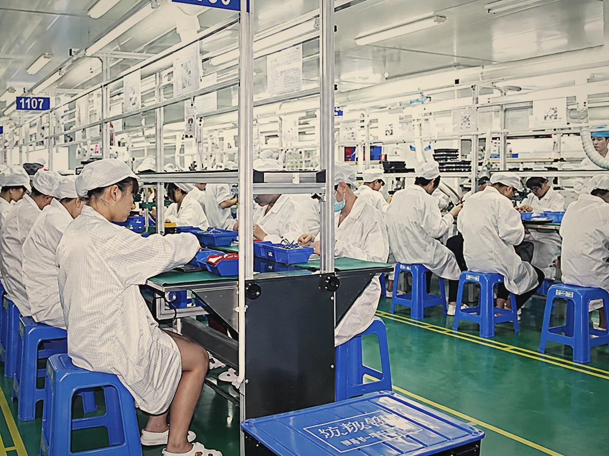 Rows of workers in white coats and caps assembling cameras at long benches on the factory floor, with blue stools and parts trays