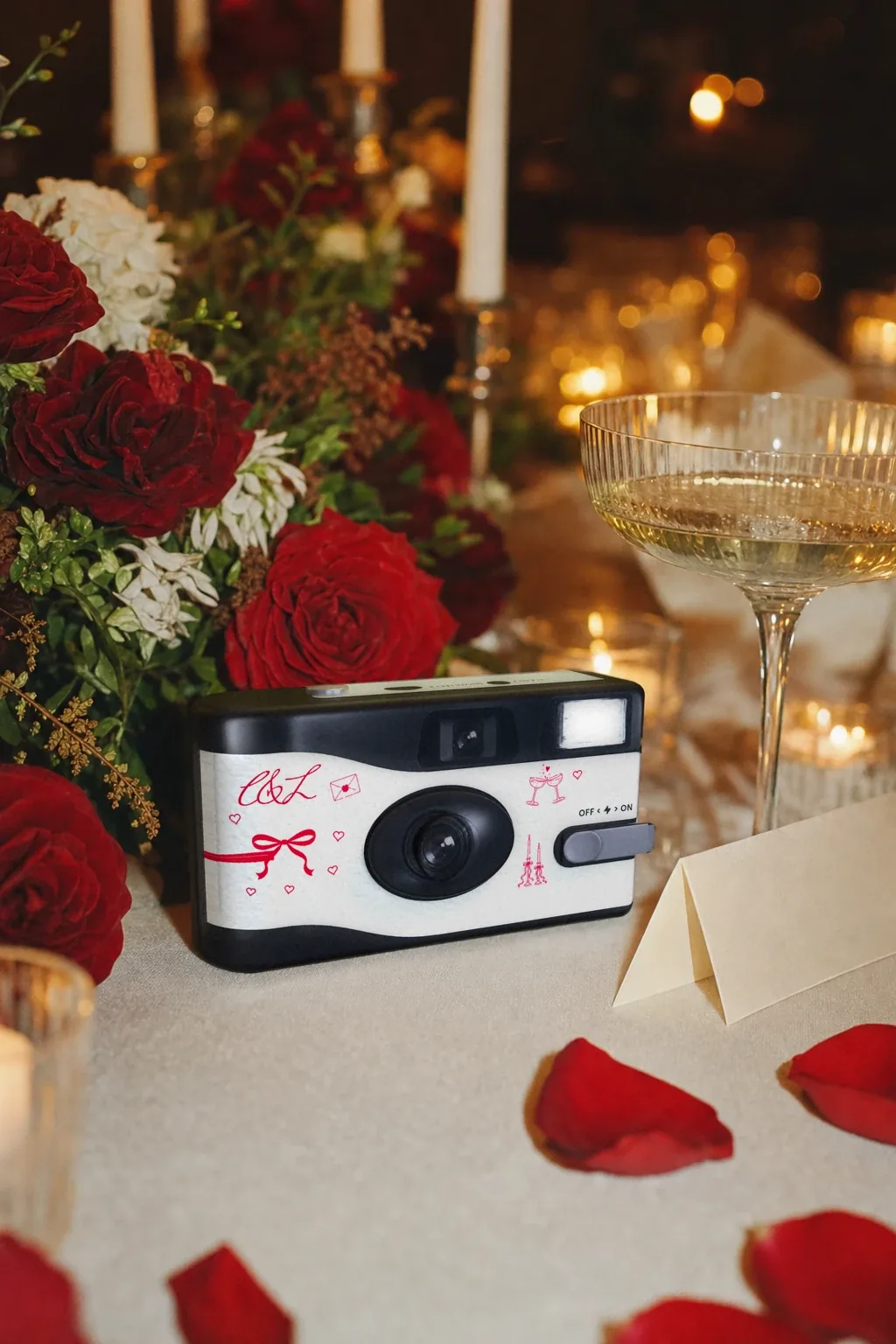 Charlotte and Liam's camera on a candlelit reception table beside deep red roses, a champagne coupe and rose petals