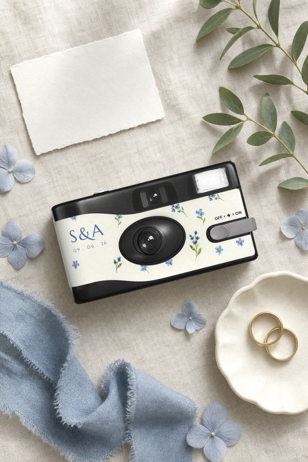 Sian and Aidan's camera laid on linen with a blank card, green leaves, blue hydrangea petals, a blue ribbon and two wedding rings in a dish